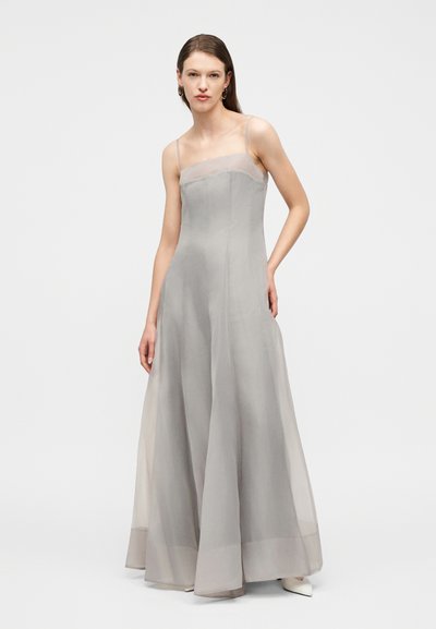 Γυναίκα με μακρύ γκρι διαφανές φόρεμα με λεπτές τιράντες, στέκεται μπροστά σε απλό λευκό φόντο.