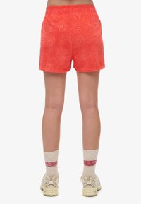 Shorts rouges texturés avec une taille élastique et des poches latérales. Portés avec des chaussettes de couleur claire et des baskets neutres, montrant un style décontracté.