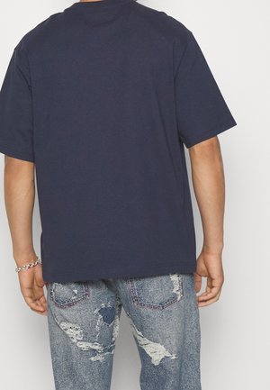 Person iført en ensfarvet mørkeblå kortærmet T-shirt og lyseblå slidte jeans set bagfra på en hvid baggrund.