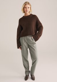 Pulover tricotat, texturat, de culoare maro, cu o croială lejeră și mâneci late, asortat cu pantaloni gri deschis, cu talie înaltă, și botine maro cu vârf ascuțit.