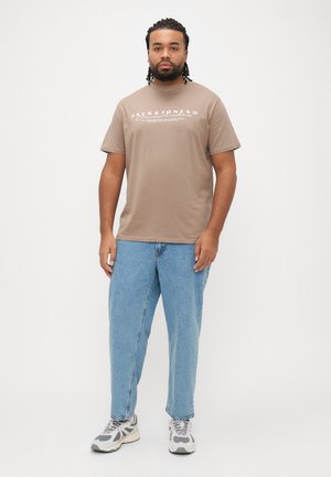 Camiseta de algodón marrón con una marca gráfica blanca, combinada con jeans de mezclilla azul claro y zapatillas grises, de pie contra un fondo blanco.