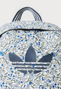 Blumenmuster-Rucksack mit weißem Grund, der blaue und gelbe Blumen zeigt. Enthält ein dunkles blaues Adidas-Logo und gestreifte Träger.