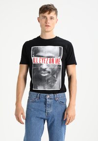 T-shirt en coton noir présentant une grande impression graphique d'un visage avec le texte "ALL EYEZ ON ME" en rouge vif au centre. Coupe classique.