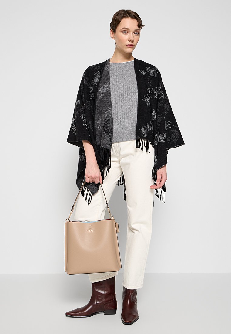 Poncho noir à franges avec motif floral gris, pull côtelé gris, pantalon crème, bottines en cuir marron et sac à main beige.