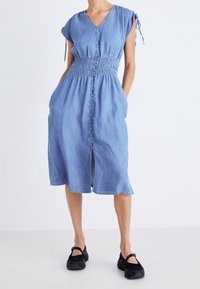 Nainen, joka on pukeutunut polvimittaiseen siniseen napitettavaan mekkoon, jossa on kerätty vyötärö ja hihattomat hihat, yhdistettynä mustiin Mary Jane -kengiin.