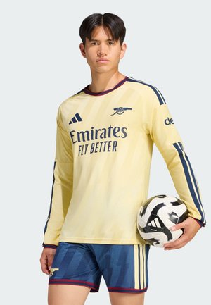 Joven con camiseta amarilla de fútbol del Arsenal y pantalones cortos azules sosteniendo un balón de fútbol Adidas blanco y negro sobre un fondo liso.