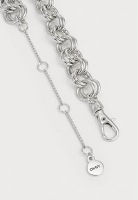 Cinturino in metallo argentato con grandi maglie, dotato di un moschettone e di un'etichetta circolare DKNY. Superficie texture e finitura lucida.