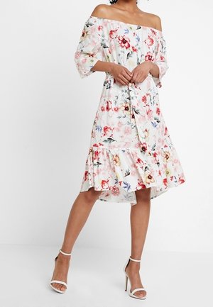 Femme portant une robe blanche épaules dénudées avec des motifs floraux roses, rouges et jaunes, associée à des sandales blanches à talons hauts.