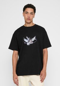 Sort bomulds T-shirt med et frontgrafik af en hvid due med udstrakte vinger, omgivet af en cirkulær linje og farvede prikker.