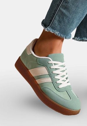 Pied portant une basket en suède verte avec des rayures blanches, des lacets blancs et une semelle marron, associée à un jean bleu à ourlet effiloché.