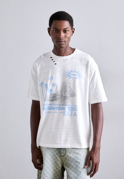Man in een witte gescheurde graphic t-shirt met blauwe tekst en afbeelding, staande tegen een effen lichtgrijze achtergrond.