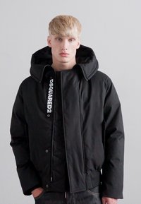 Young man with short blonde hair wearing a black Dsquared2 hooded jacket, standing against a plain light gray wall.