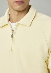 Homme portant un sweat à col zippé jaune pâle sur une chemise blanche, montrant le col zippé et un logo subtil sur la poitrine.