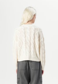 Suéter de malha grossa em creme com padrão de cabos, bainha canelada e mangas largas. Usado com calças cargo cinzas, visto de costas.