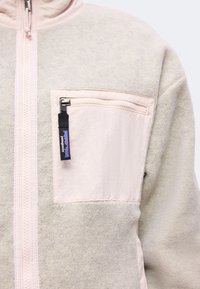 Κοντινή λήψη μπεζ fleece μπουφάν με ανοιχτό ροζ τσεπάκι στο στήθος με φερμουάρ και μαύρο τράβηγμα φερμουάρ με λογότυπο Patagonia.