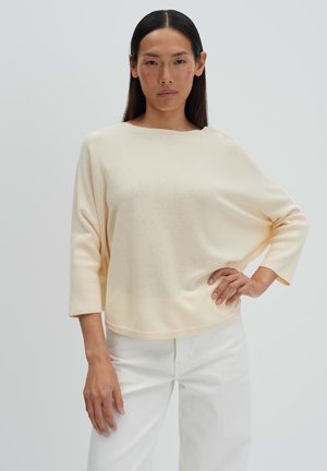 Woman with straight black hair wears loose cream sweater with 3/4 sleeves and white pants, standing with one hand on hip against plain background.