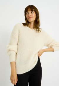 Suéter de punto beige claro con ajuste holgado, mangas largas y puños acanalados; textura suave y dobladillo ligeramente redondeado, combinado con pantalones negros.