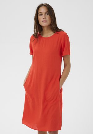 Žena s dlhými hnedými vlasmi, oblečená v jasne oranžových krátkych šatách s okrúhlym výstrihom a rukami v predných vreckách, stojaca na bielom pozadí.