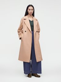 Femme aux longs cheveux noirs portant un manteau beige, un haut vert foncé, un jean large bleu et des bottes marron, debout devant un fond blanc uni.