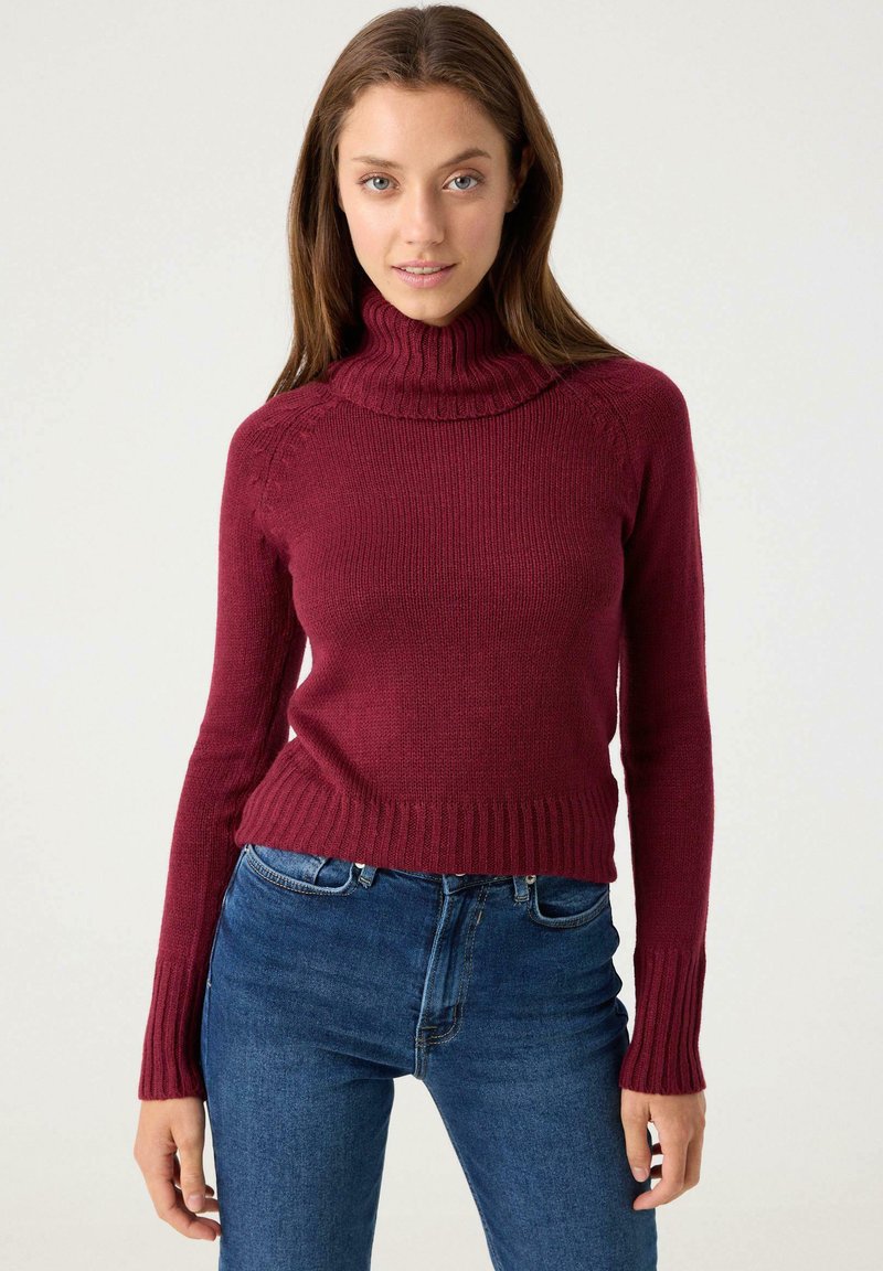 Suéter de cuello alto de punto burdeos con dobladillo y puños acanalados, que presenta un diseño ajustado y mangas largas, combinado con jeans azules.