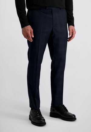 Homme portant un pantalon habillé bleu marine sur mesure avec un pli, des chaussures de ville noires et une chemise noire à manches longues, se tenant sur un fond blanc.