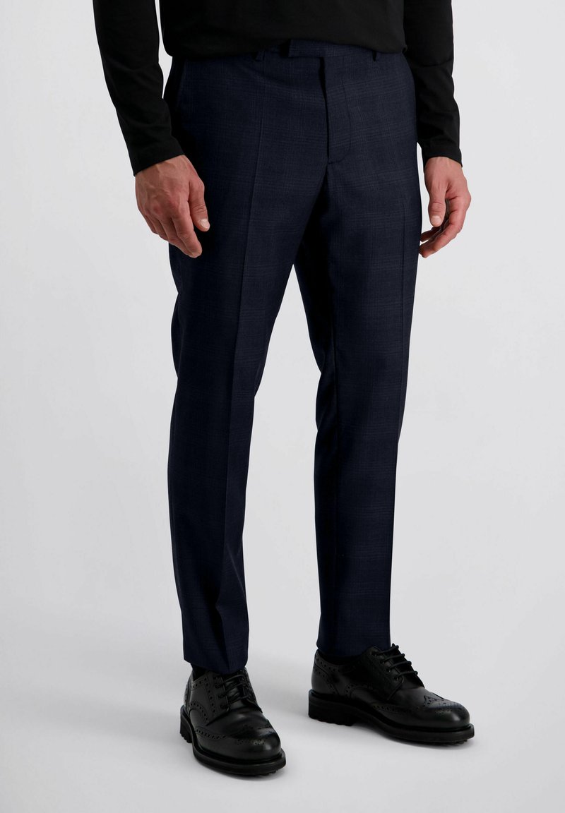 Man in op maat gemaakte donkerblauwe dress-pantalon met plooi, zwarte dress-schoenen en zwart langemouwenshirt op een witte achtergrond.