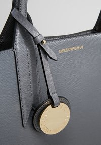 Szara, teksturowana skórzana torba na ramię z okrągłą, złotą zawieszką z logo, z ozdobnym detalem w postaci kokardy na uchwycie oraz wytłoczonym nazwiskiem marki.