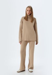 Maglione beige a collo alto in maglia, con vestibilità comoda, abbinato a pantaloni a sigaretta coordinati. Tessuto morbido, design casual e scarpe bianche.