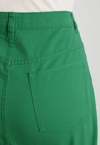 Pantalones cortos de mezclilla verdes que cuentan con un bolsillo trasero y detalle de costura visible. La tela parece suave con un ligero brillo.