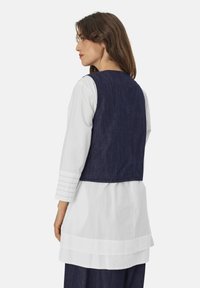 Denimweste mit tailliertem Rücken, getragen über einer weißen Bluse mit Lagen und gepunkteten ärmeln. Schlichtes Design, dunkelblaue Farbe und glatte Textur.