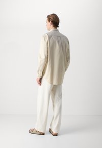 Beige langarmshirt met een ontspannen pasvorm, enkele knoopmanchetten, achterpand met inzetstuk, gecombineerd met losse witte broek en beige sandalen.