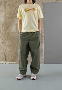 Camiseta de algodón amarilla clara con un gráfico logo de Nike en naranja, combinada con pantalones holgados de color verde oliva y zapatillas blancas con detalles rojos.