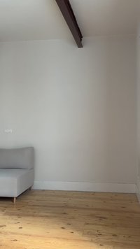 Γκρι καναπές απέναντι σε λευκό τοίχο με σκούρα δοκό οροφής και ανοιχτόχρωμο ξύλινο πάτωμα σε ένα άδειο δωμάτιο.