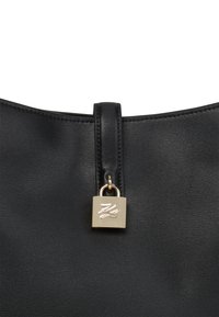 Melnas ādas soma ar zelta kvadrātu slēdzeni, kas pievienota vertikālai ādas siksnai centrā.