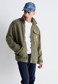 Chaqueta de felpa verde oliva con cremallera y un alto cuello, dos bolsillos laterales y un bolsillo en el pecho. Usada sobre una camisa blanca y combinada con unos vaqueros.