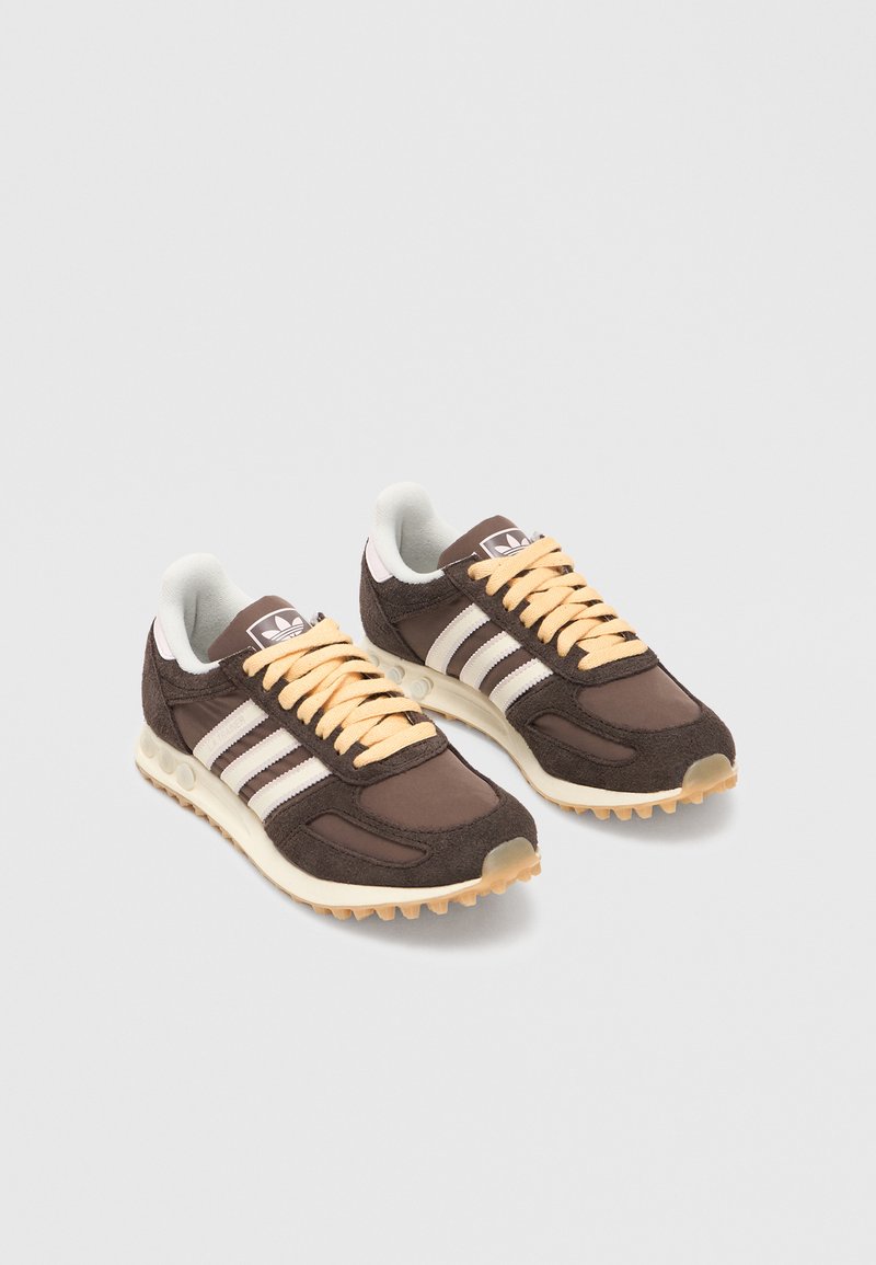 Pár hnědých tenisek Adidas s bílými pruhy a béžovými tkaničkami na bílém pozadí, natočeno doprava.