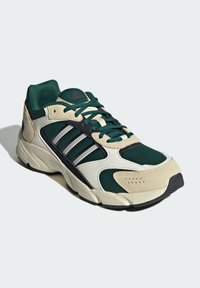 Chaussure de sport verte et beige en matériaux en maille et en suède, avec des accents noirs, trois rayures argentées et une semelle texturée.