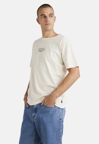 Cream-colored cotton t-shirt with short sleeves and a round neckline. Features a logo graphic on the chest and a textured finish.