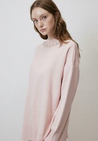 Pulover roz cu guler cerc, având un accent de perlă la decolteu. Creat dintr-un material moale, cu mâneci lungi și o croială lejeră.