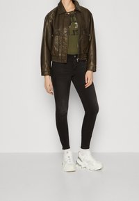Chaqueta bomber de imitación piel marrón con bolsillos frontales, una camiseta verde con estampado debajo, combinada con jeans skinny negros y zapatillas deportivas blancas gruesas.