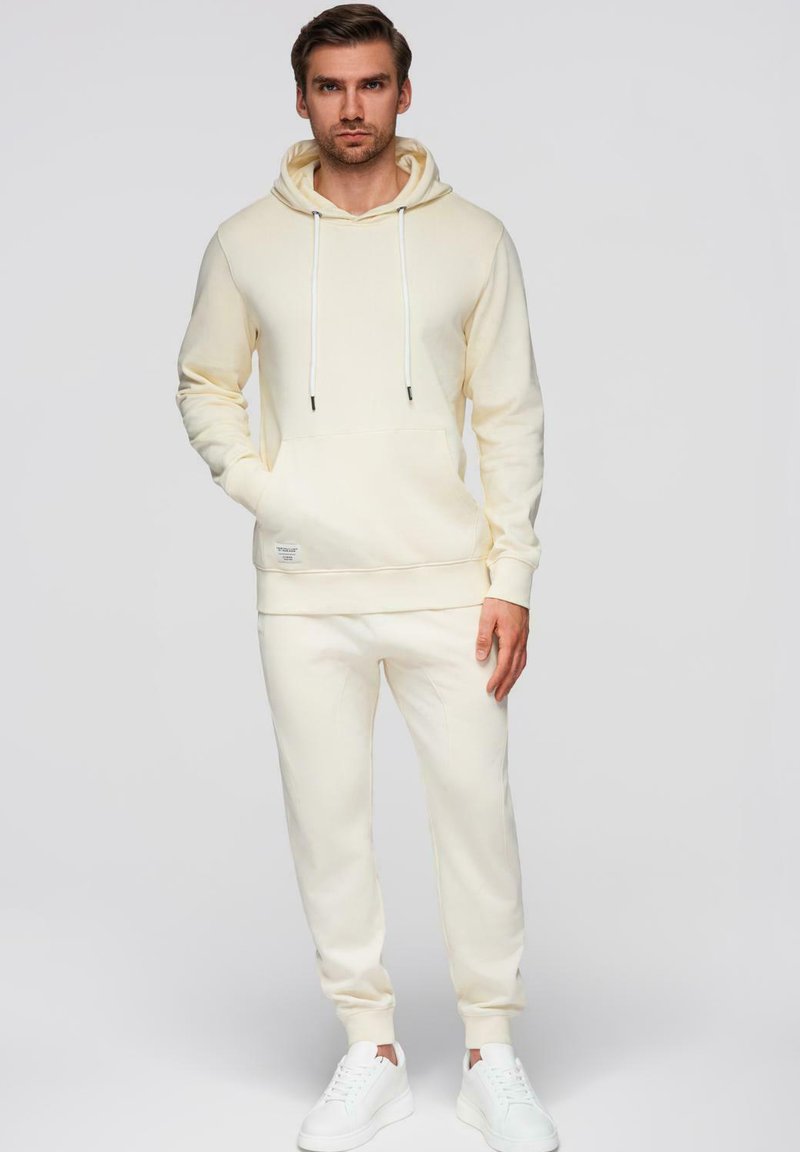 Homme portant un sweat à capuche crème et un pantalon de survêtement assorti, debout avec une main dans la poche, sur un fond uni.