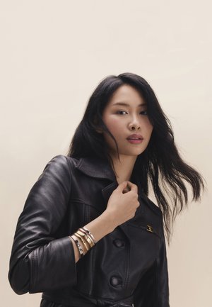 Joven con cabello largo y negro que lleva una chaqueta de cuero negra y múltiples pulseras de oro y plata en la muñeca, con un fondo liso.