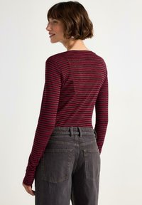 Langärmliges Oberteil mit roten und schwarzen Streifen, aus weichem Stoff. Kombiniert mit hoch geschnittenen, dunkelgrauen Jeans, die über Gesäßtaschen und einen taillierten Bund verfügen.