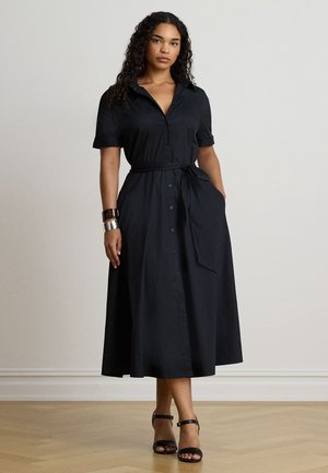 Une femme porte une robe midi noire à manches courtes, boutonnée sur le devant et nouée à la taille, ainsi que des sandales noires, sur un fond de studio uni.