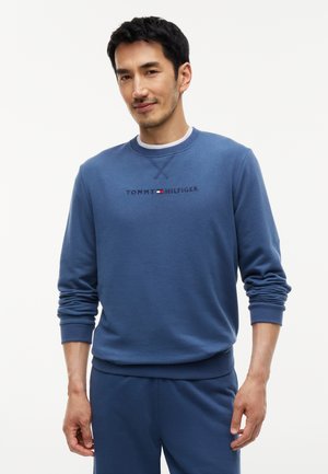 Άνδρας που φορά μπλε φούτερ Tommy Hilfiger και κατάλληλο παντελόνι, στέκεται μπροστά σε έναν απλό λευκό φόντο.