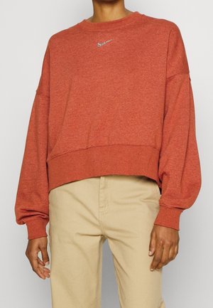 Une personne portant un sweat-shirt oversize couleur rouille avec un petit logo Nike argenté et un pantalon beige, se tenant contre un fond uni.