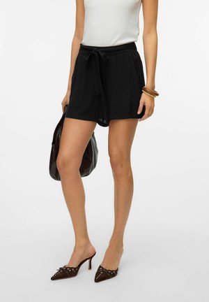 Shorts noirs avec une taille à nouer, ornés de broderies en dentelle. Associés à un haut blanc et un sac marron. Chaussures marron à bout pointu avec des embellissements.