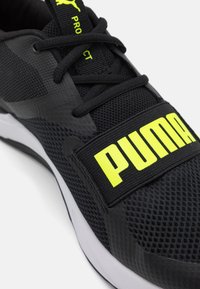 Pantof dintr-un material athletic negru, cu o parte superioară din plasă, talpă din cauciuc și o curea proeminentă galbenă cu logo-ul "PUMA" pe mijlocul piciorului. Accente texturate și șireturi.