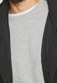 Szara bluza z miękką tkaniną i okrągłym dekoltem, częściowo zakryta czarną kurtką z gładkiego, lekko błyszczącego materiału z zamkami błyskawicznymi.