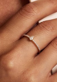 Gouden ring met een delicate band voorzien van een kleine peervormige diamant omringd door kleinere stenen, geplaatst op een hand met textuur in de huid.